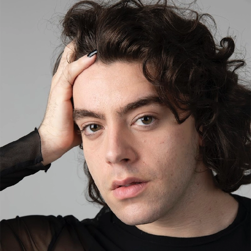 A person with wavy brown hair and fair skin looks into the camera, resting their left hand on their head. They wear a sheer black long-sleeved top. Their expression is calm and thoughtful, and the background is plain light gray.