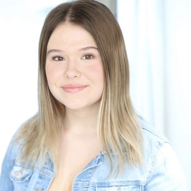 Erin Gerasimovich, a young woman with straight, light brown hair fading to blonde at the ends, smiles softly. She wears a light blue denim jacket over a light top, posed in front of a bright, softly blurred background.