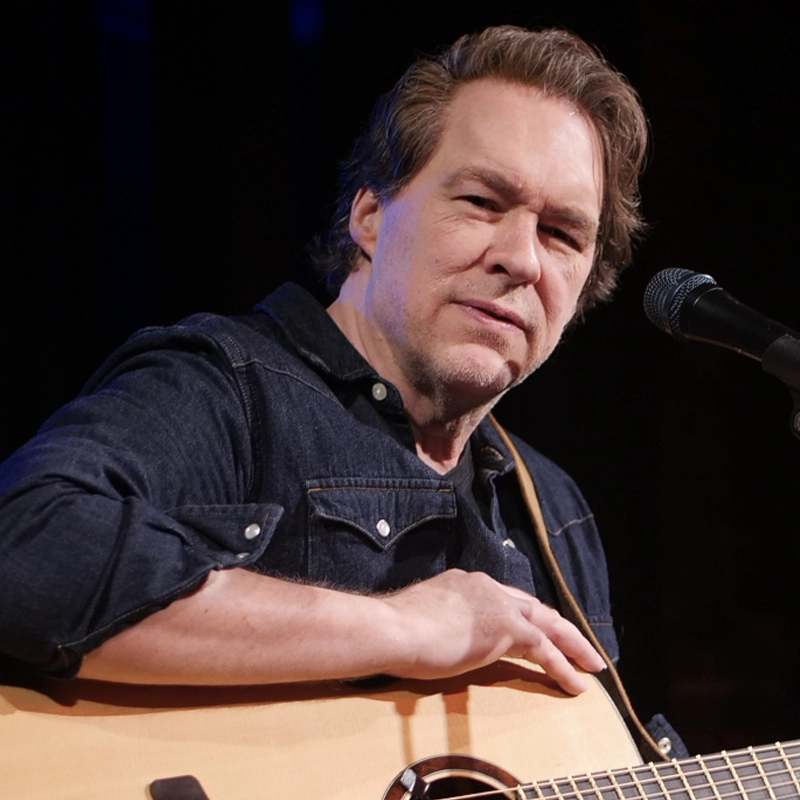 A man with light skin and brown hair, wearing a dark denim shirt, sits with an acoustic guitar. He appears to be playing or preparing to play and looks thoughtfully toward the camera. A microphone on a stand is visible in front of him against a dark background.