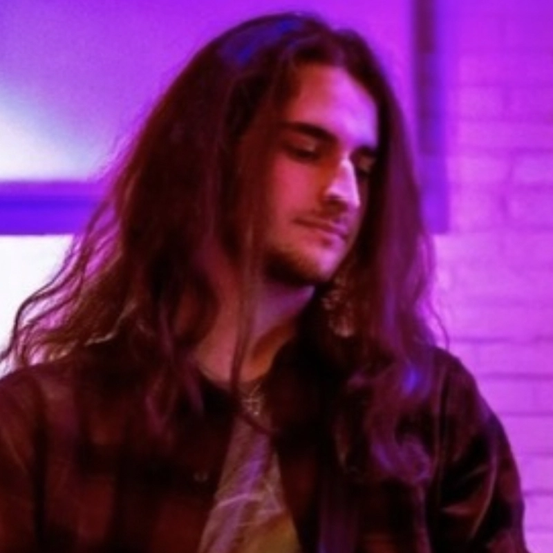 Jonathan Goldberg, a young man with long brown hair and light skin, looks downward with a calm expression. He wears a dark plaid shirt, standing against a purple-lit background of white bricks that creates a soft, artistic mood.
