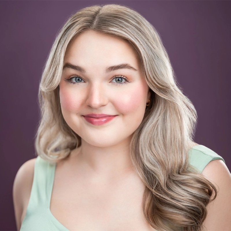 A young woman, Olivia Goodman, with long, wavy blonde hair wears a light green sleeveless top. She has fair skin, blue eyes, and rosy cheeks, and is smiling gently. The background is a plain, deep purple color with soft, even lighting.
