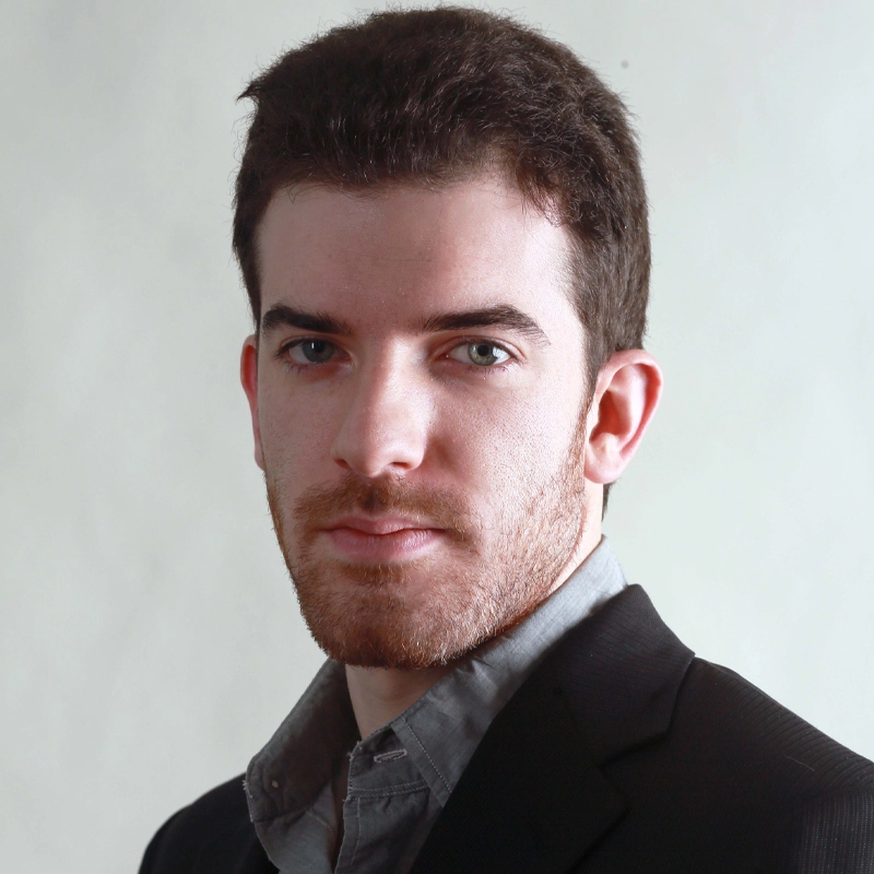 A young man with short brown hair, a trimmed beard, and blue eyes looks at the camera. He is wearing a dark suit jacket over a light grey collared shirt. The background is plain and light, giving the portrait a professional appearance.