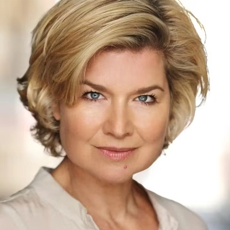 Shinnerie Jackson, a woman with short, layered blonde hair and blue eyes, looks directly at the camera, smiling softly. She wears a light beige top, while the softly blurred background in light tones brings focus to her face.