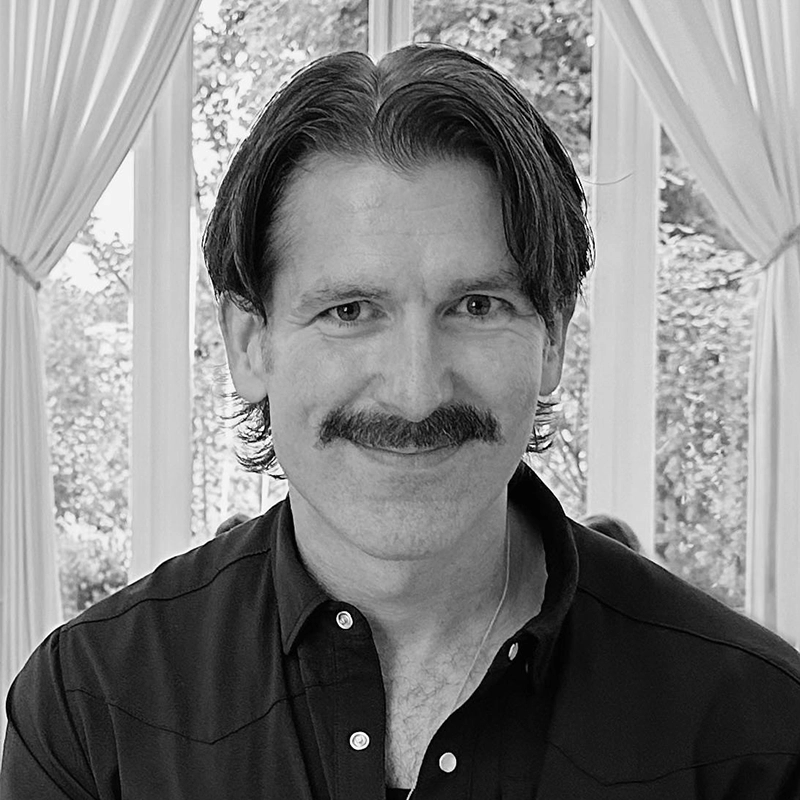 A man with medium-length dark hair and a thick mustache smiles at the camera. He wears a dark button-up shirt. Light streams through tall windows and white curtains behind him, with trees visible outside in this Kellie Ishmael black and white photo.