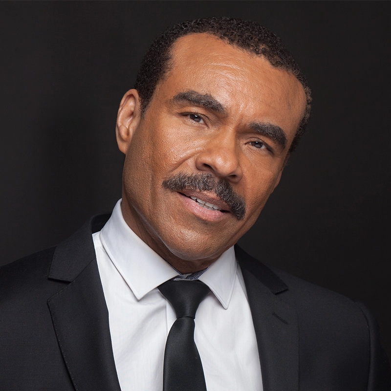 A man with short curly hair, a mustache, and medium brown skin wears a black suit, white shirt, and black tie. Reminiscent of Joseph Joubert’s thoughtful demeanor, he tilts his head slightly and smiles softly before a plain dark background.