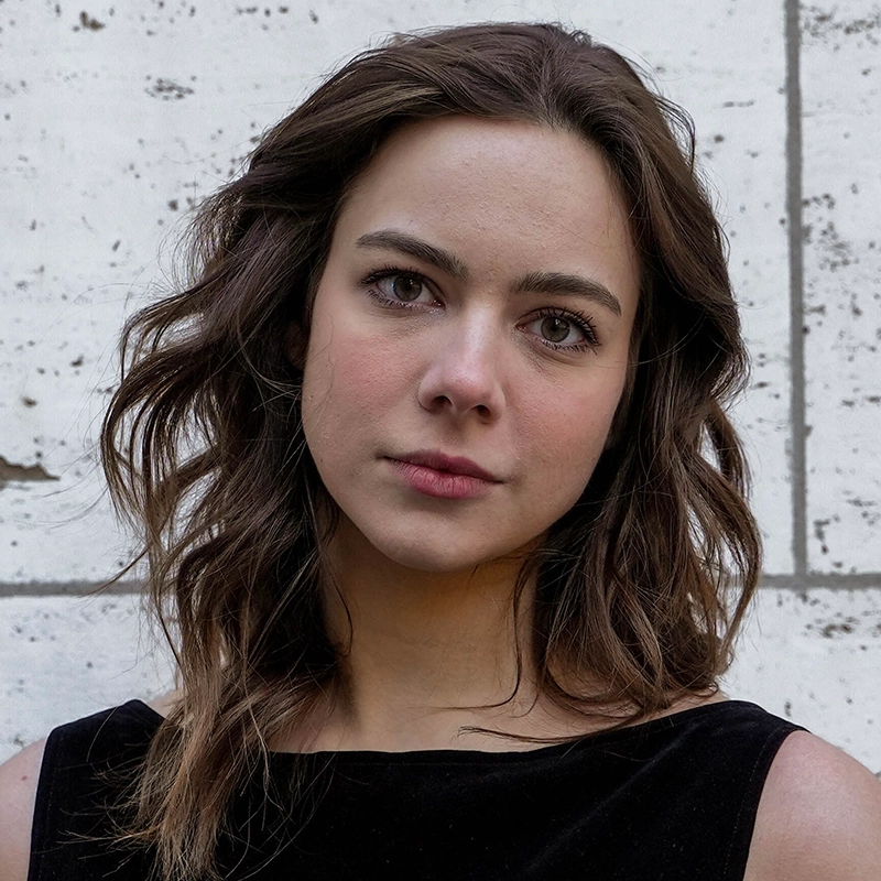 Karenna Peterson, a young woman with wavy brown hair and fair skin, gazes at the camera with a neutral expression. She wears a sleeveless black top and stands before a lightly textured, off-white stone wall with visible lines and markings.