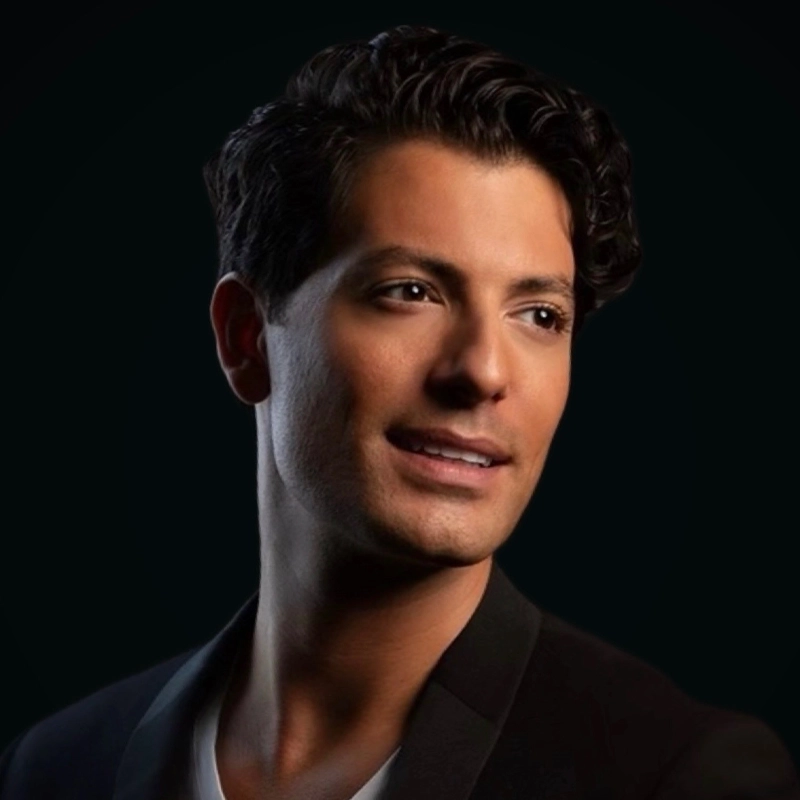 Jonathan Karrant, with wavy dark hair and smooth skin, looks slightly to the side, smiling softly. He wears a dark blazer over a white shirt. The black background makes his face and upper body stand out clearly.
