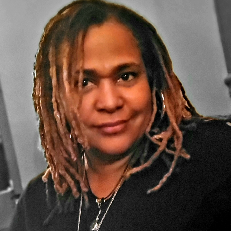Kellie Ishmael, with light brown, shoulder-length locs, wears a black top and a necklace. She faces the camera with a gentle smile, her warm skin tone and highlighted hair contrasting against the blurred gray background.