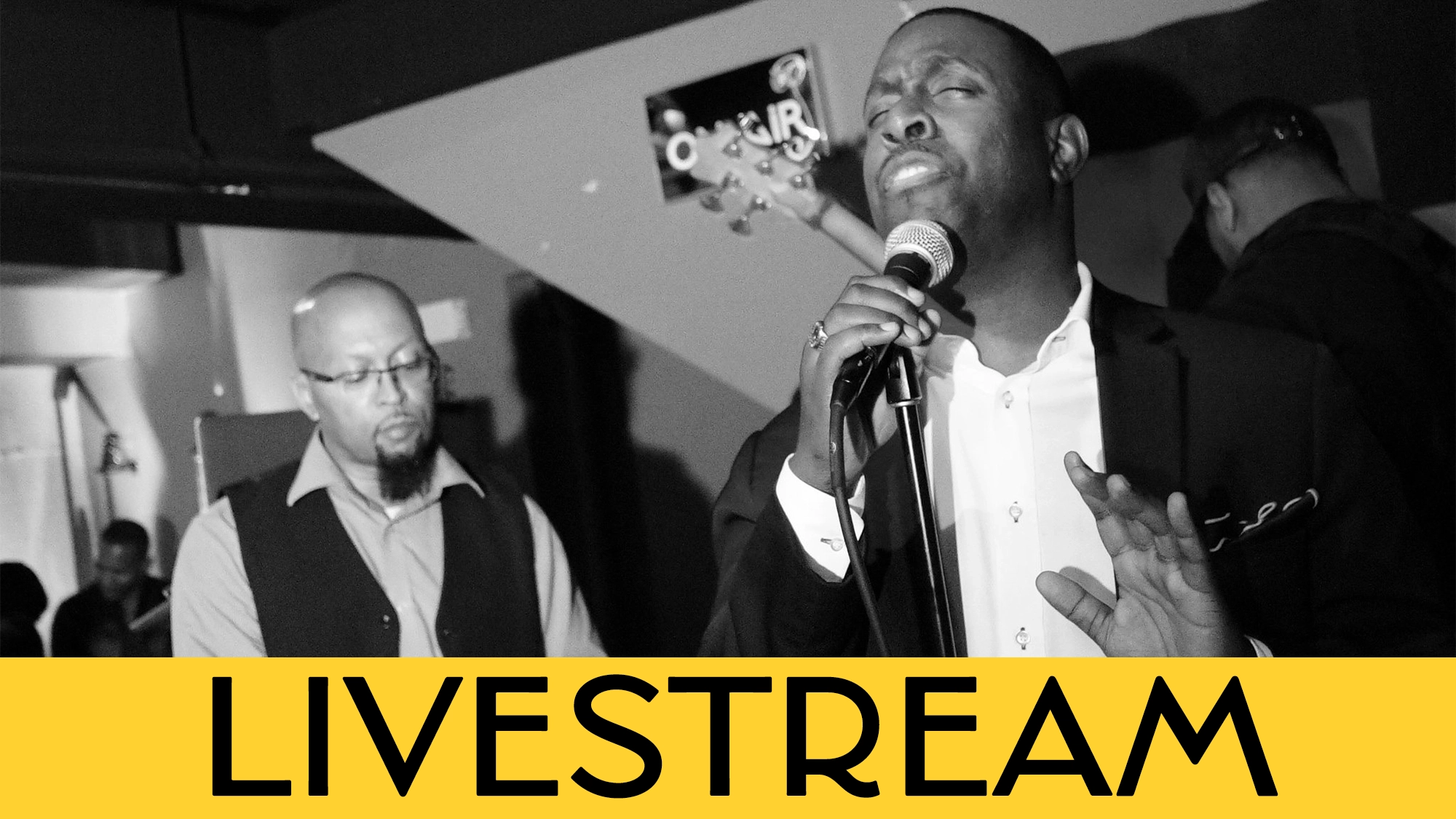 Black and white photo of Ken Alston Jr passionately singing into a microphone, eyes closed, with another man playing keyboard behind him. Both wear formal attire. A yellow banner at the bottom displays “LIVESTREAM” in bold black letters.