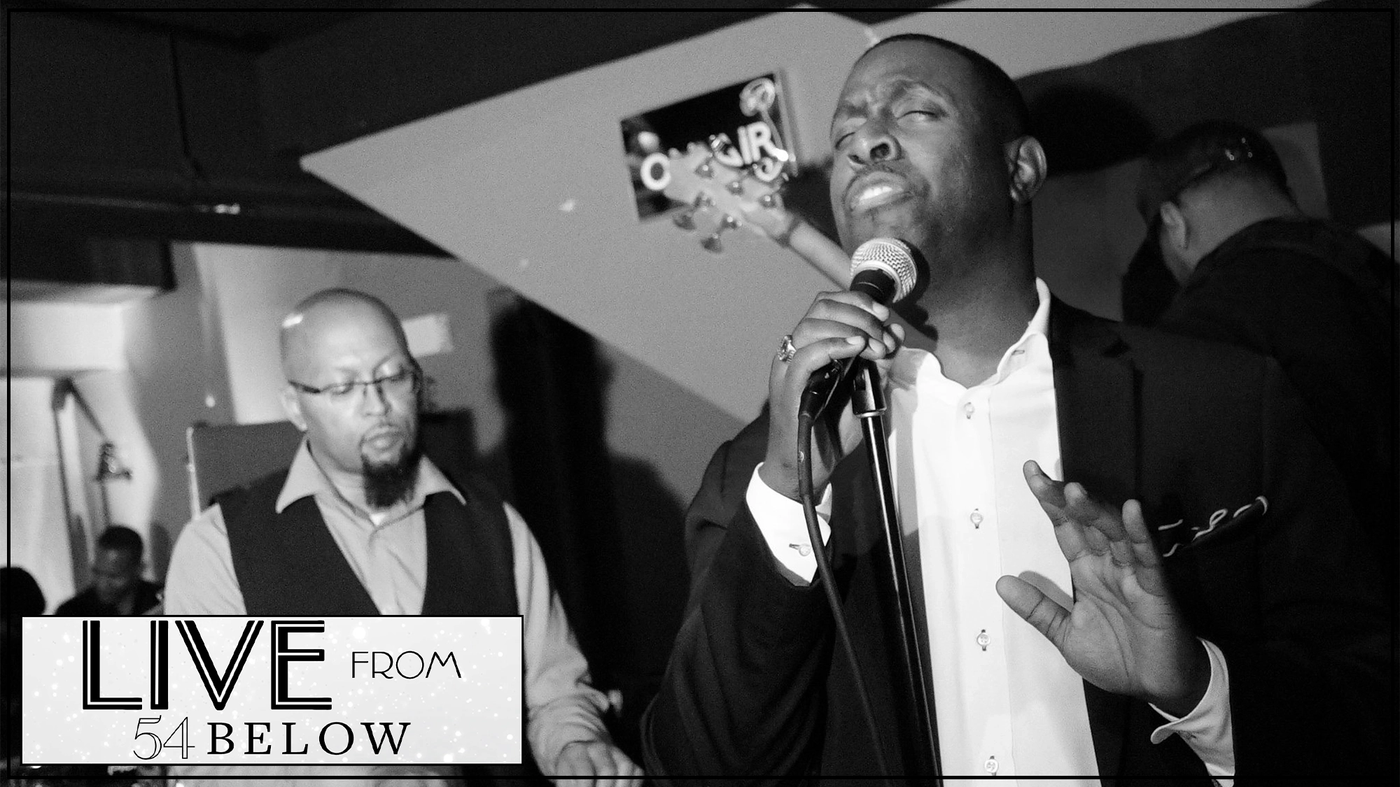 Black and white photo of Ken Alston Jr in a suit performing passionately into a microphone, eyes closed and hand raised. A musician in glasses plays keyboard behind him. Text in the corner reads 