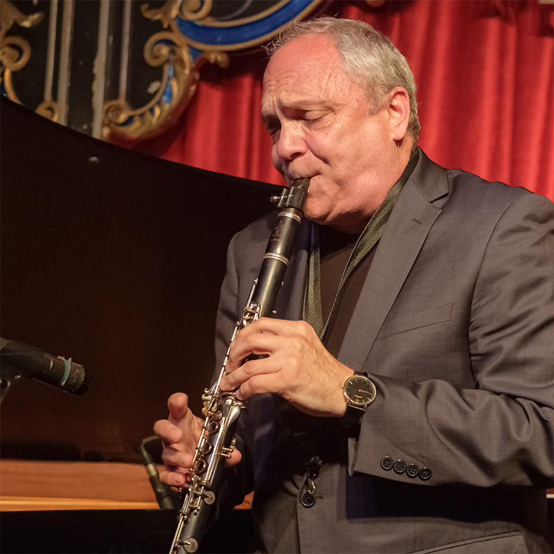 Renowned clarinetist Ken Peplowski, in a suit, plays passionately on stage before a piano, red curtains, and ornate decorations. Eyes closed in deep focus, he performs with a microphone set before him.