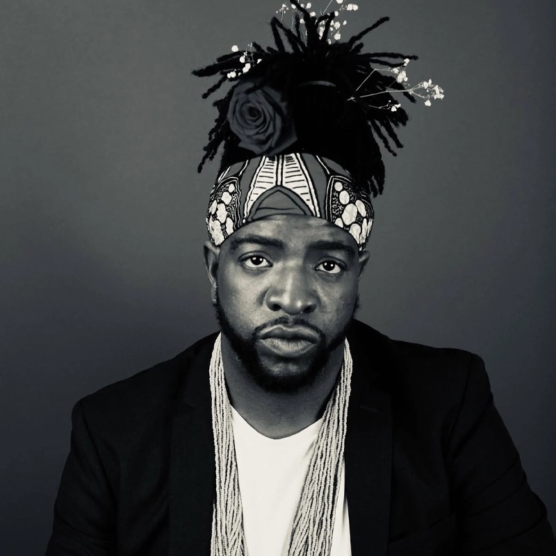 A Black man with a serious expression wears a dark blazer, white shirt, and layered beaded necklaces. His hair is styled in locs with a patterned headband, decorated with a large rose and small flowers on top. The background is plain and monochromatic.