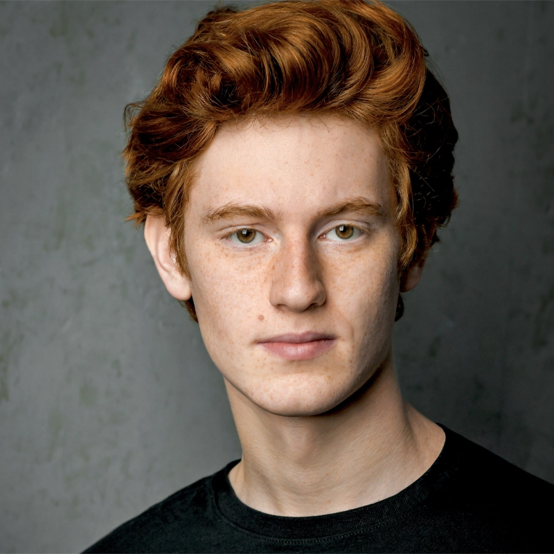 A young person with fair skin, short curly red hair, and freckles looks directly at the camera with a neutral expression. They are wearing a black shirt and standing against a plain gray background.