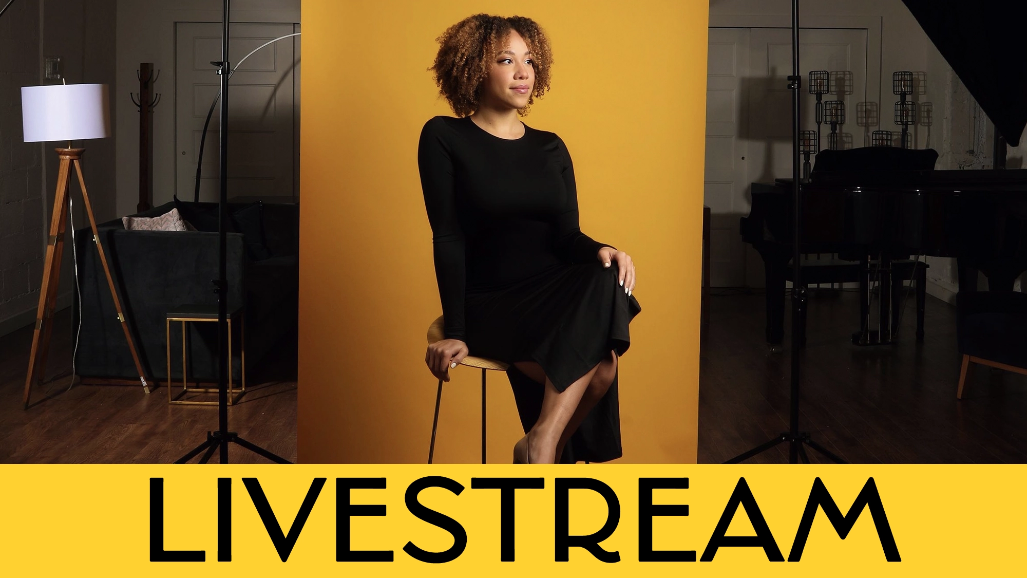 A woman with curly hair in a long black dress sits on a stool in front of a yellow backdrop. Sidney Nicole Wilson is barefoot, one leg crossed, as she prepares for her 
