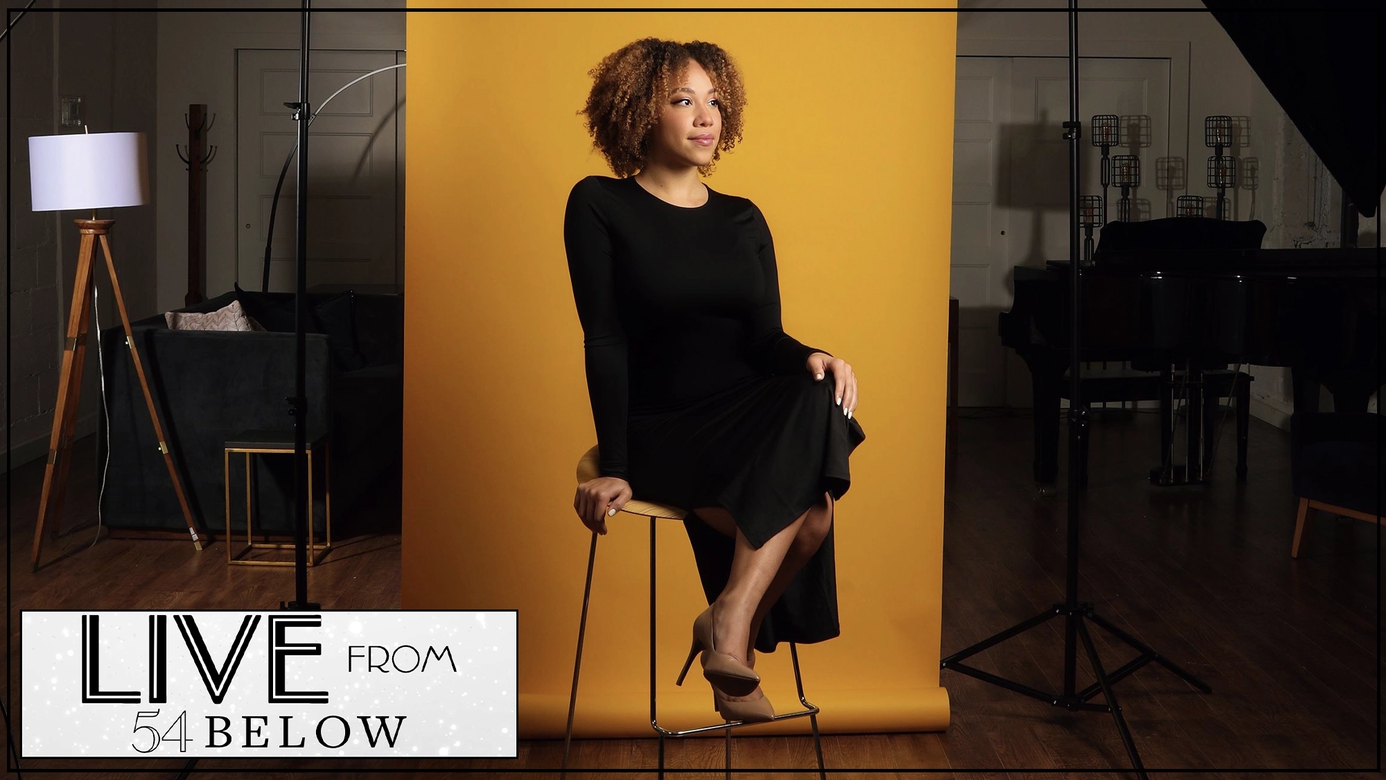 LIVE Sidney Nicole Wilson A woman with curly hair in a long black dress sits on a stool in front of a yellow backdrop. The performance space features a piano, lamp, and couch. A sign reads “LIVE FROM 54 BELOW,” as Sidney Nicole Wilson prepares for the Livestream.