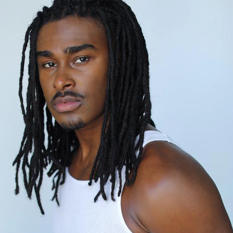 A young Black man, Lamont Walker II, with long black dreadlocks and a thin goatee gazes confidently at the camera. He wears a sleeveless white shirt that reveals his shoulder and upper arm, set against a neutral light blue background.