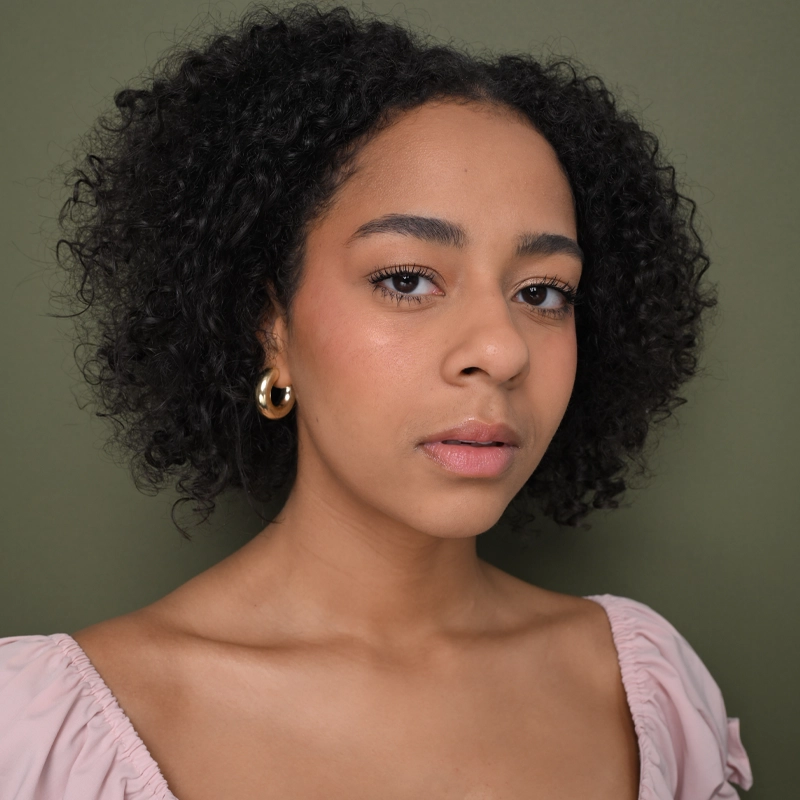 Kiara Michelle Lee, a woman with short curly hair, stands against an olive green background, wearing a light pink top with puffed sleeves and gold hoop earrings. She gazes softly at the camera with a neutral expression.