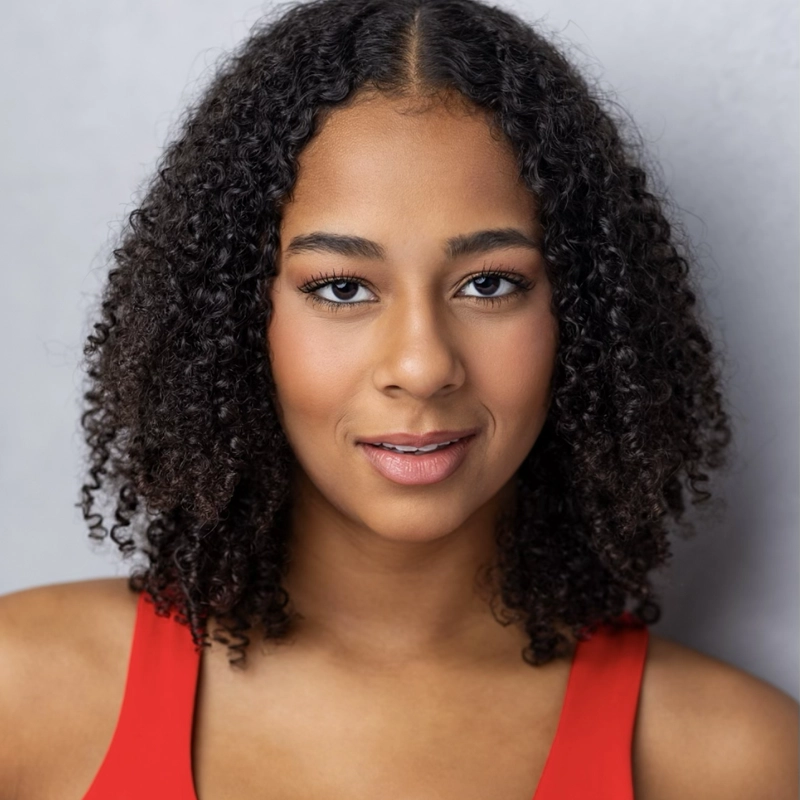 A young woman with medium brown skin and curly, shoulder-length black hair smiles softly. She is wearing a sleeveless red top and stands in front of a plain light gray background. Her expression is calm and confident.