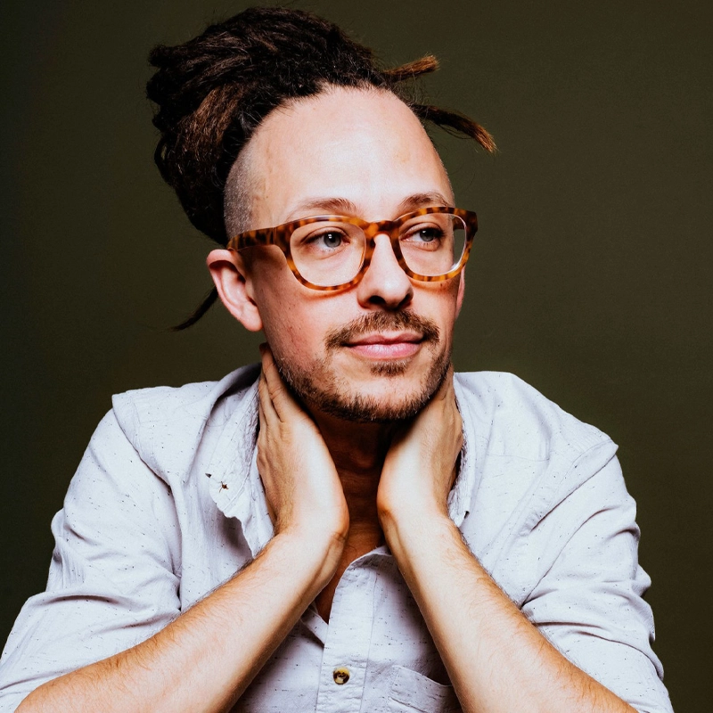 A person with light skin, wearing tortoiseshell glasses and a white shirt, rests their hands on their neck. They have Philippe Lemm-inspired dreadlocked hair tied up with shaved sides, a mustache, and a goatee. The background is plain olive green.