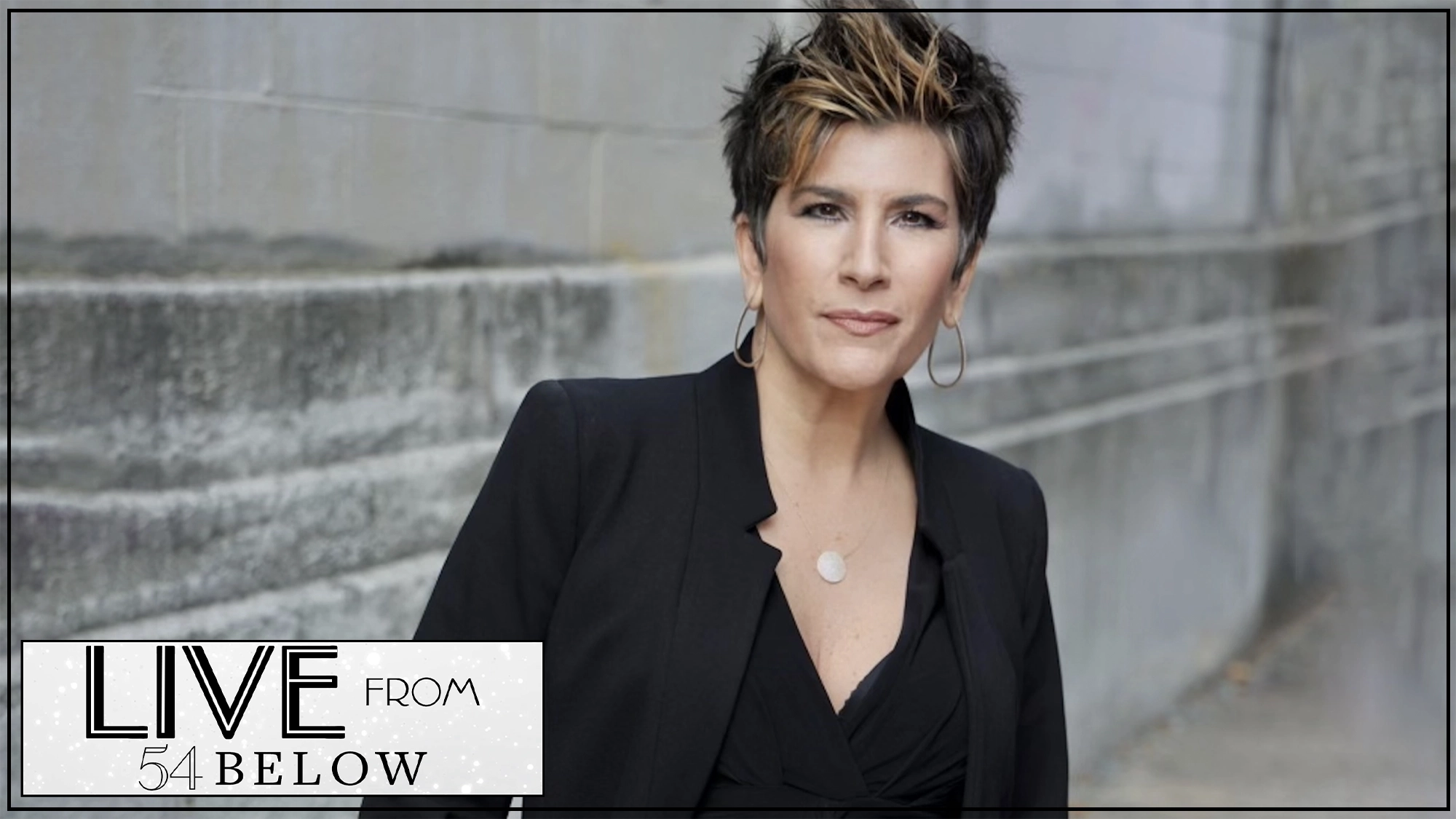 A person with short, spiked brown hair and hoop earrings, dressed in a black blazer and v-neck top, stands against a gray concrete wall. In the corner, a graphic reads 