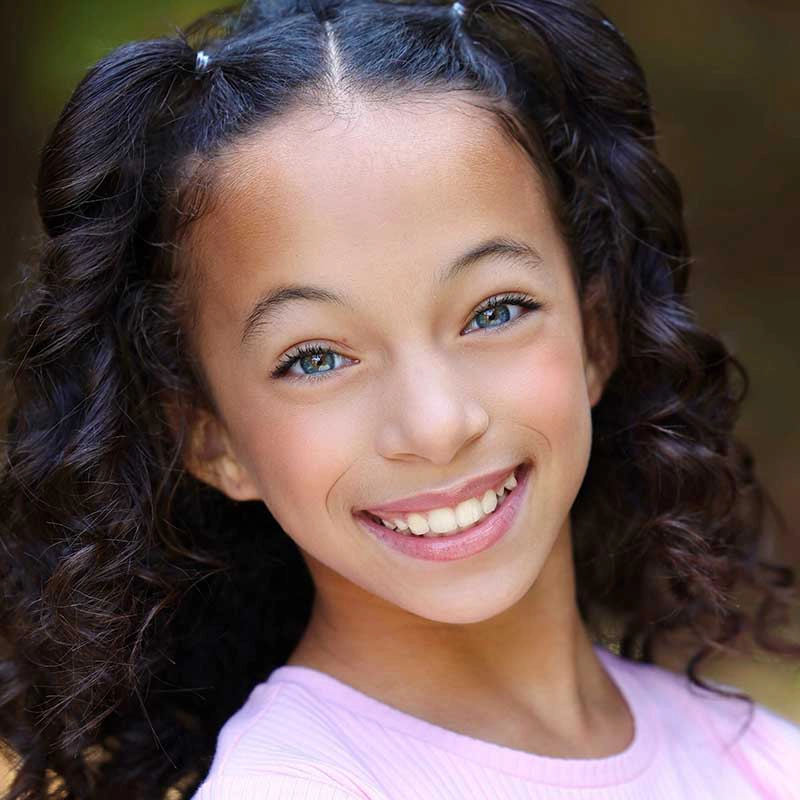 Lianne Gomes, a young girl with light brown skin, curly dark hair in pigtails, and blue-green eyes, smiles brightly. She wears a light pink shirt and stands outdoors with a blurred natural background.