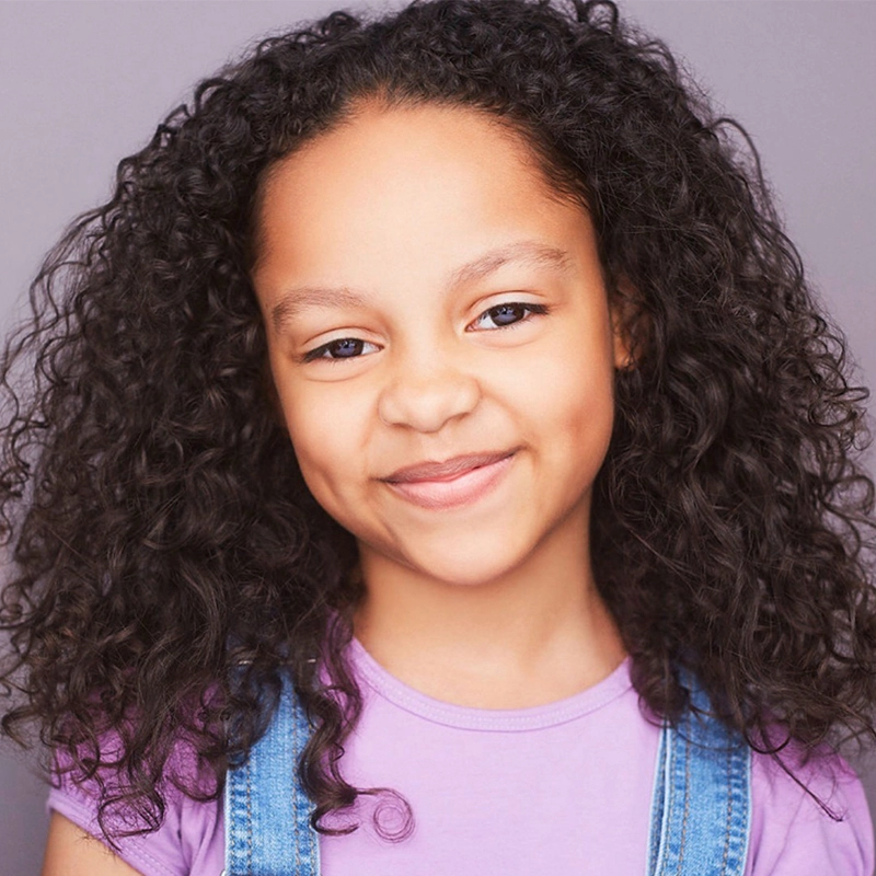 A young girl with long, curly dark hair smiles softly at the camera. McKenzie S. Lewis wears a light purple t-shirt and blue denim overalls. The plain, light gray background highlights her friendly, expressive face.