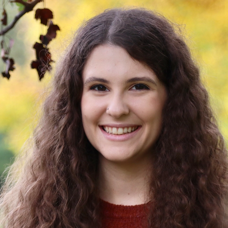A young woman with long, thick, curly brown hair smiles warmly. Megan Smythe wears a rust-colored sweater and has a nose ring. The background is outdoors, with blurred yellow-green foliage and a branch in the upper left corner.