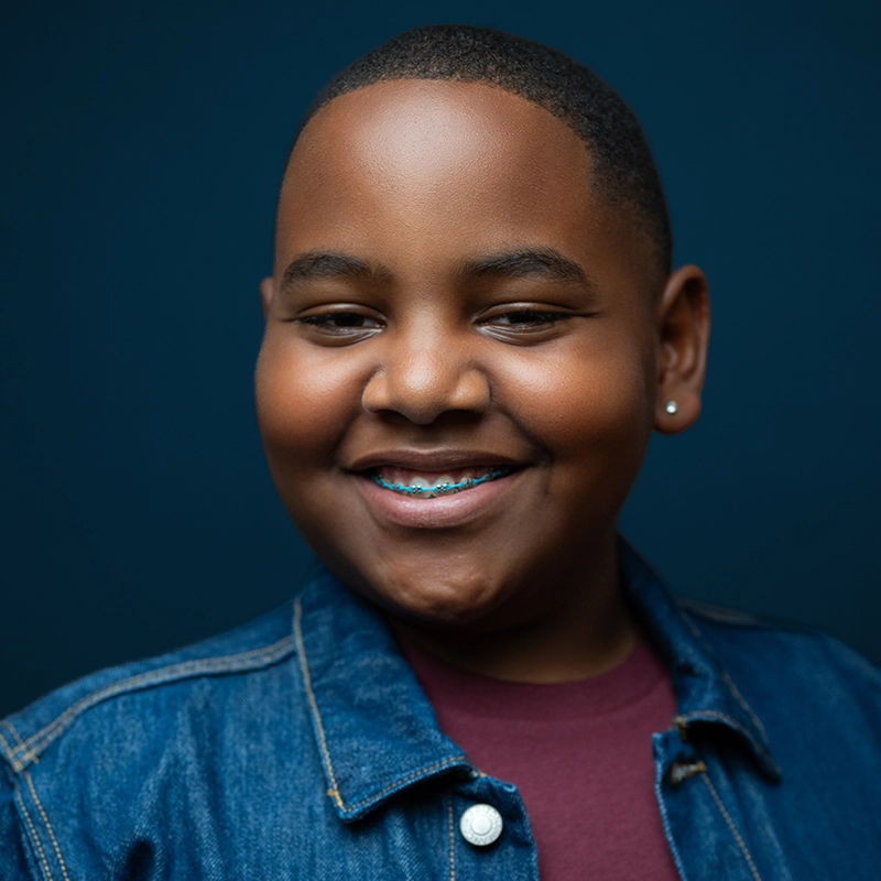 Mekhi Richardson, a smiling child with short hair and braces, wears a denim jacket over a maroon shirt. An earring in the left ear glints as Mekhi stands against a solid dark blue background, the lighting highlighting a joyful expression.
