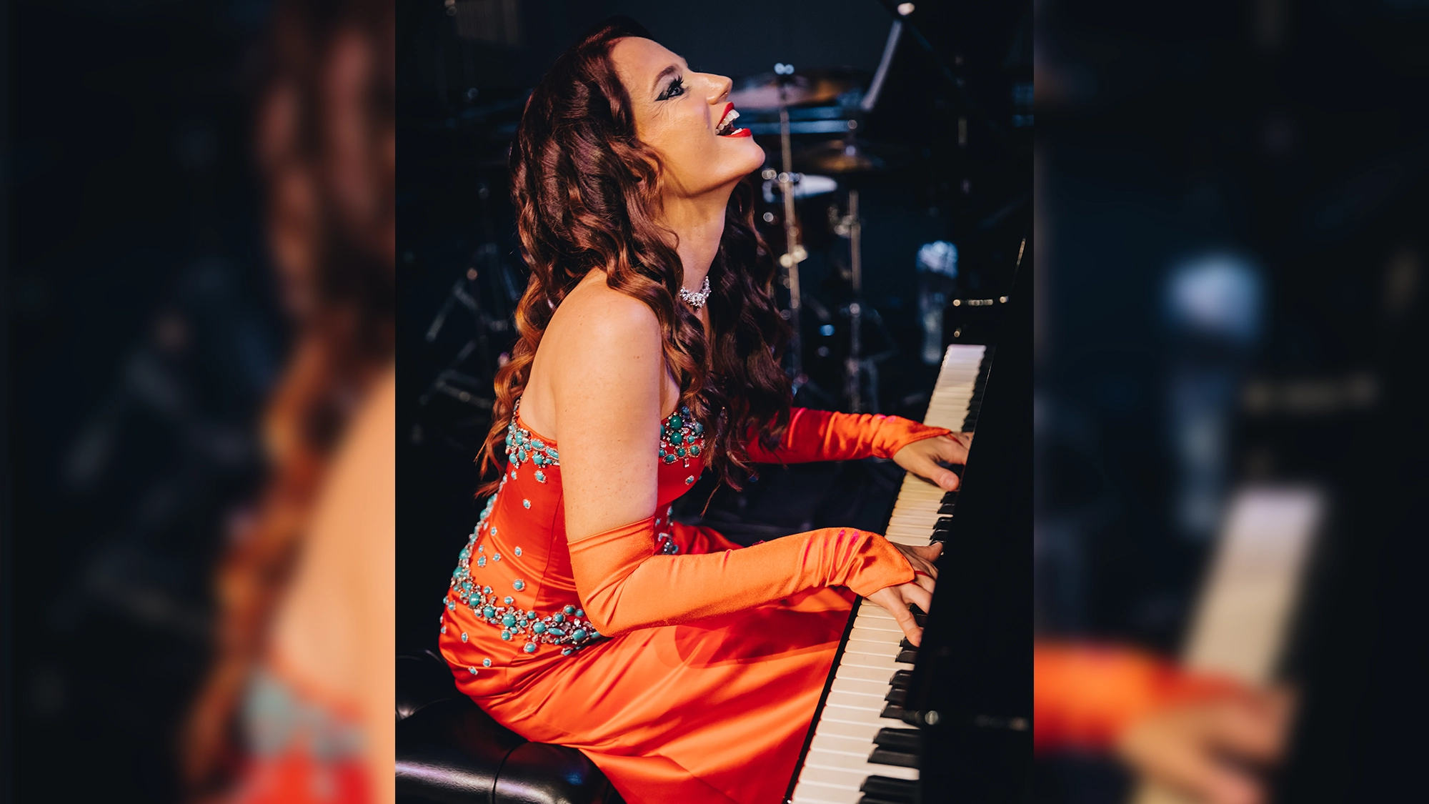 Mia Vassilev site image 26 Mia Vassilev, in a sparkling orange gown and long matching gloves, plays a piano for Keys on Fire, smiling and looking upward. She has long wavy brown hair, a jeweled necklace, and the dimly lit background shows blurred musical equipment.