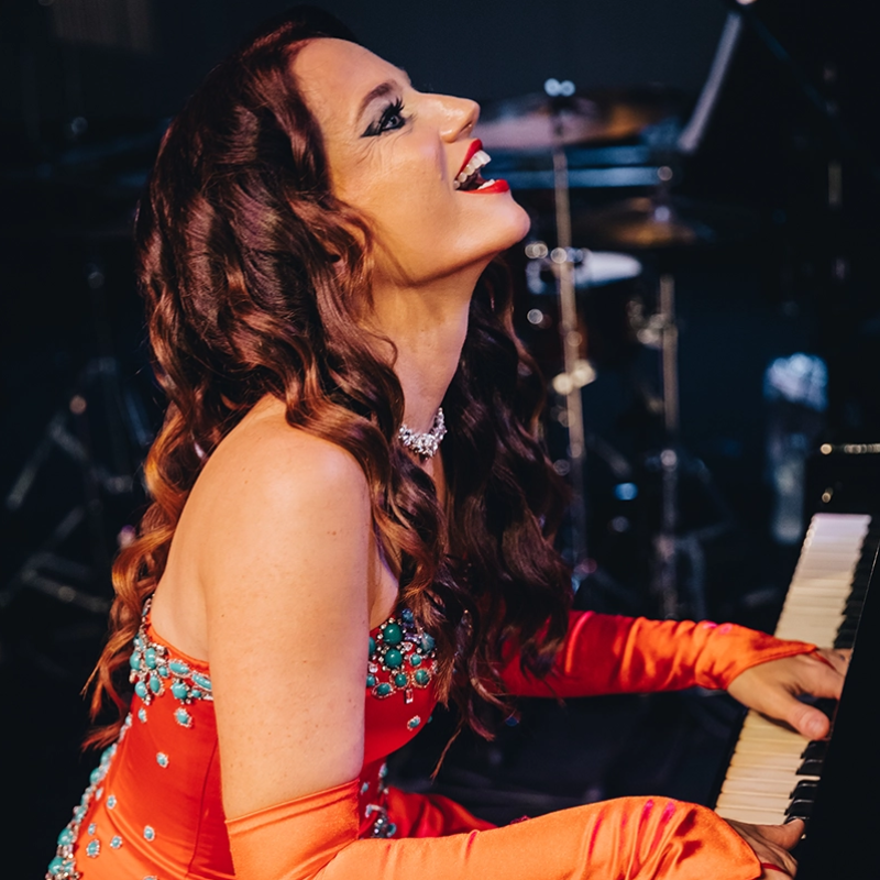 A woman with long, wavy auburn hair, wearing a jeweled red dress and gloves, laughs joyfully while playing a piano. She has dramatic makeup and a sparkling necklace. Musical instruments and stage equipment are blurred in the background.