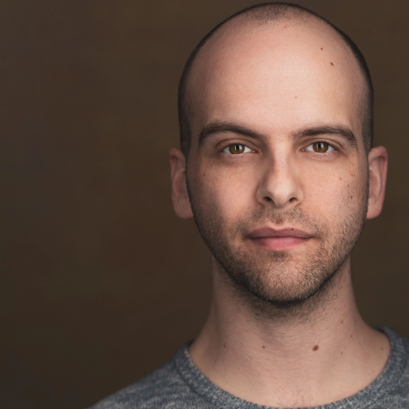 A young bald man with fair skin and stubble, Cameron Moser, looks directly at the camera with a neutral expression. He wears a gray sweater against a warm brown background, with soft, even lighting highlighting his facial features.