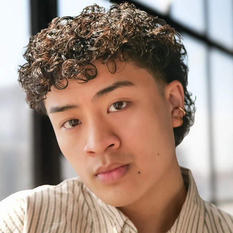 A young person with short, curly dark hair and a light brown shirt looks directly at the camera. They have smooth skin, a mole above their upper lip, and wear a gold hoop earring. Sunlight streams through large windows in the background.