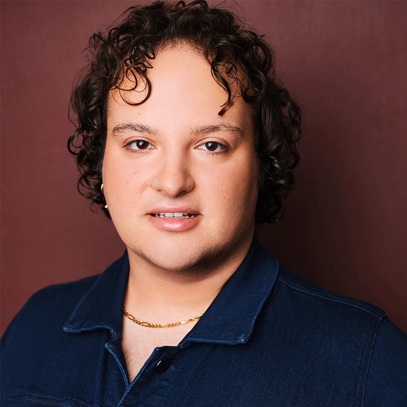 Nicholas Milan, with curly brown hair, light skin, and brown eyes, smiles softly. He wears a dark blue collared shirt, a gold chain necklace, and a small gold hoop earring against a solid burgundy background.