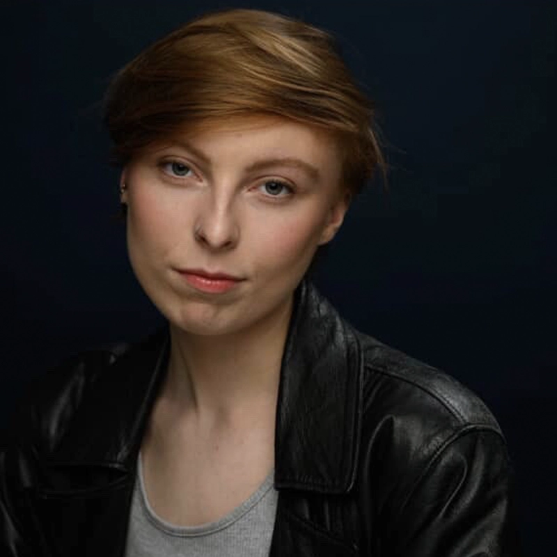 A person with short, light brown hair and fair skin wears a black leather jacket over a gray shirt. They look directly at the camera with a neutral expression. The background is dark, highlighting their face and upper body.