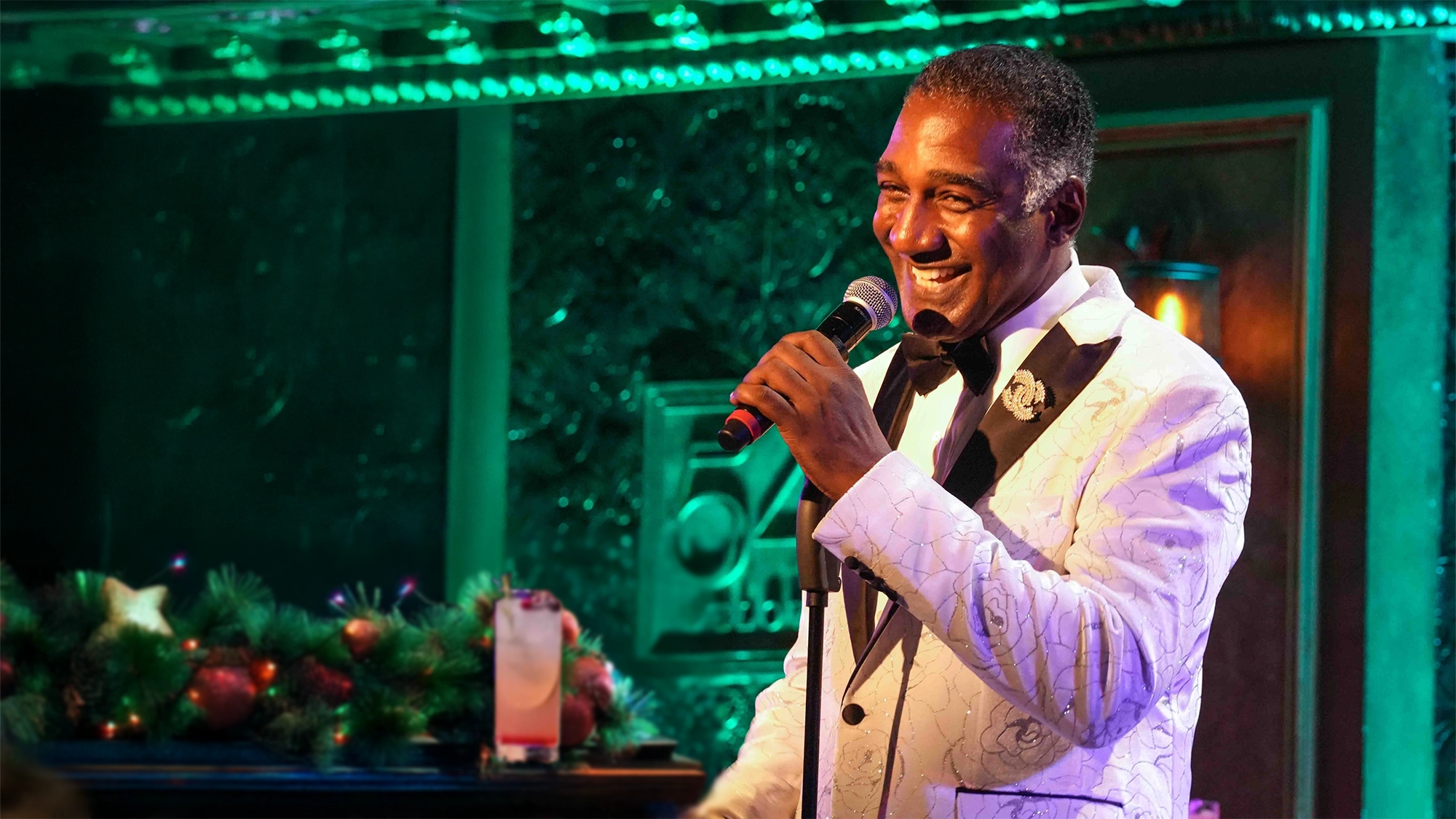 Norm Lewis site image 25 A smiling Norm Lewis in a white floral tuxedo jacket holds a microphone, performing onstage at Christmas Lives Here. Festive garland and a drink on the piano set the scene, with green and turquoise lights creating a lively Christmas atmosphere.