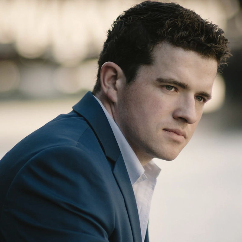 Emmett O'Hanlon, a man with short, dark curly hair and light skin, wears a blue suit jacket over a white shirt. He looks thoughtfully to the right with a neutral expression; the background is softly blurred with light tones.