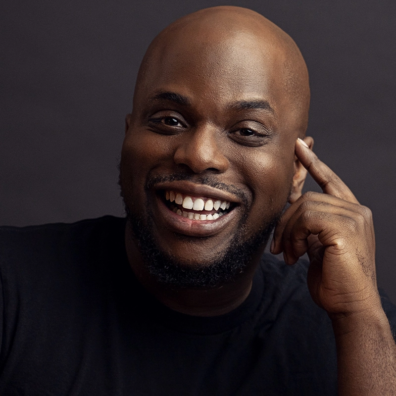 A smiling bald Black man, Patrick Dailey, with a neatly trimmed beard wears a black shirt. He is looking at the camera, showing his teeth, and touching his right temple with his index finger. The background is dark and simple.