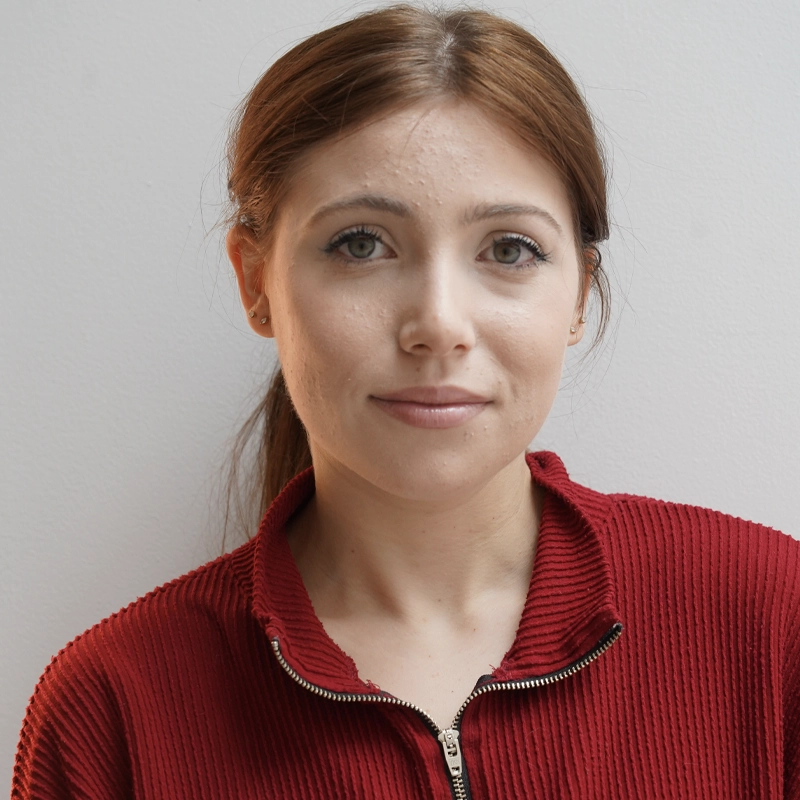 A young woman with light skin and straight brown hair tied back, wearing a textured red zip-up sweater. She has light eyes, neutral makeup, and a calm expression. She stands in front of a plain, light-colored wall.