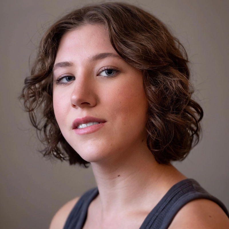 Young person with short, wavy brown hair and light skin looks at the camera with a neutral, slight smile. They wear a sleeveless dark top against a plain, muted background. Lighting is soft, highlighting natural facial features.