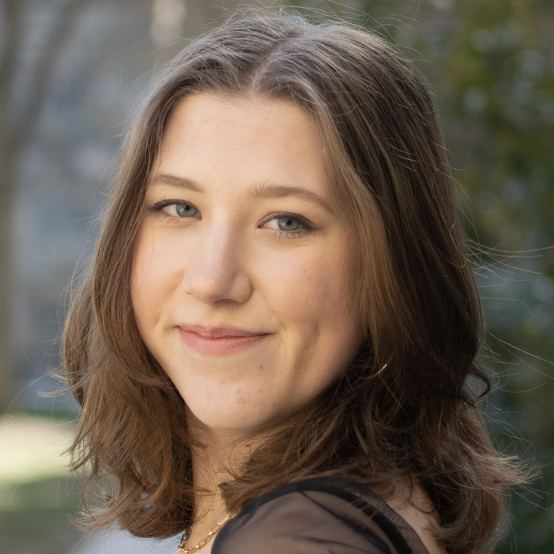 A young woman with light skin and shoulder-length brown hair smiles gently at the camera. She has blue eyes and wears subtle makeup. She is outdoors, with a blurred background of greenery and soft natural light.