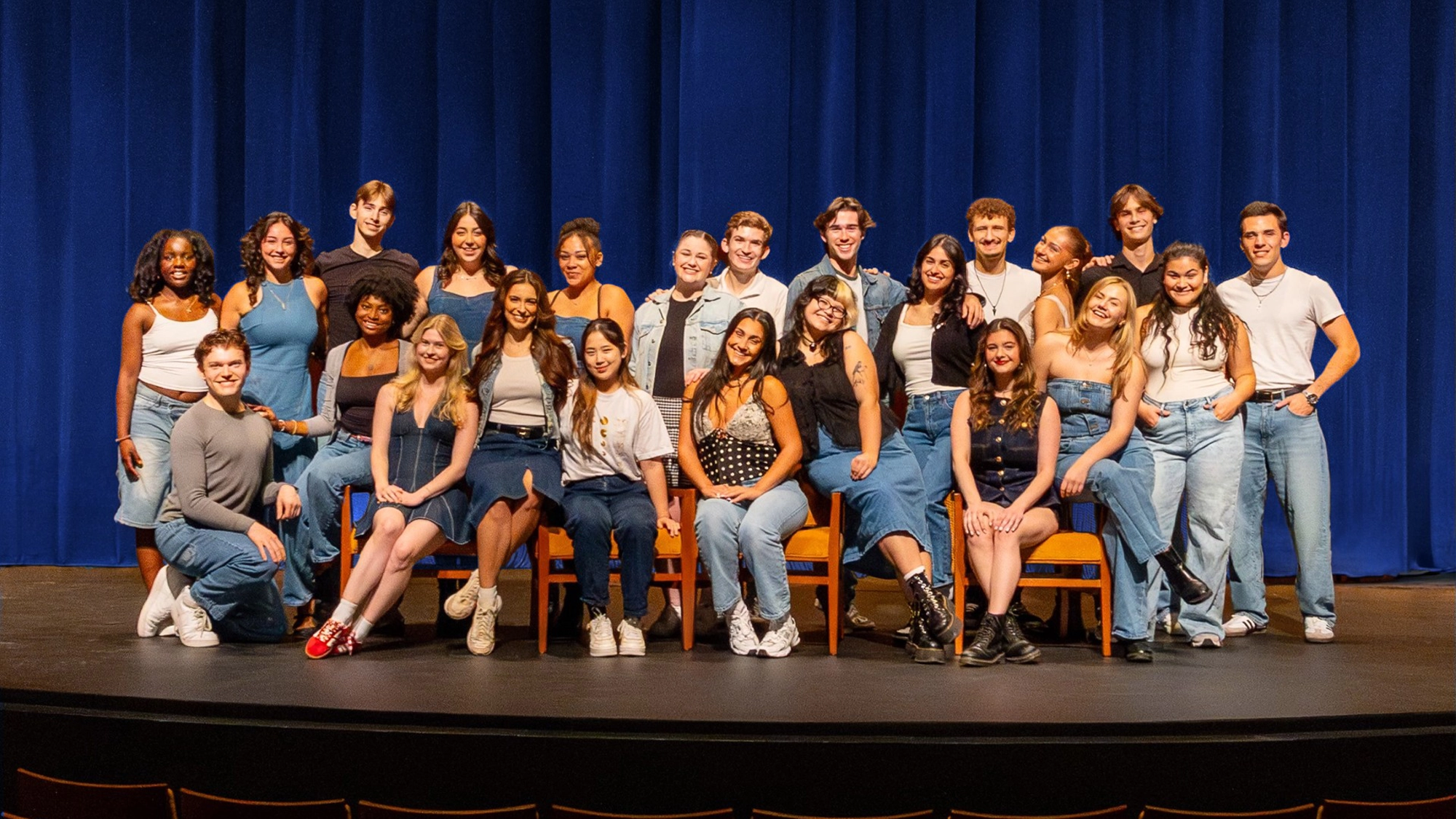 A diverse group of 22 young adults from the BFA Musical Theatre Class of 2026 at Point Park University poses on stage in front of blue curtains. Most wear denim or white tops. The mood is cheerful and casual, with some seated and others standing in a well-lit space.