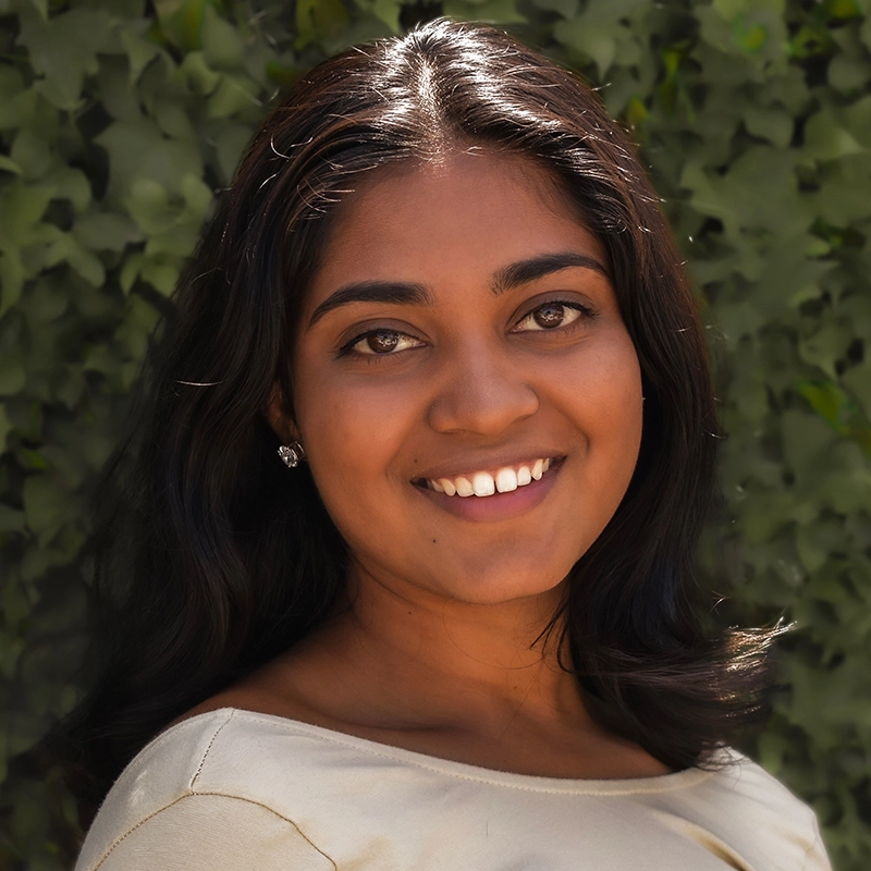 Camilla Lockhart, a woman with medium brown skin and long dark hair, smiles warmly at the camera. She wears a light-colored top and stud earrings, with green leafy foliage in the background suggesting an outdoor setting.