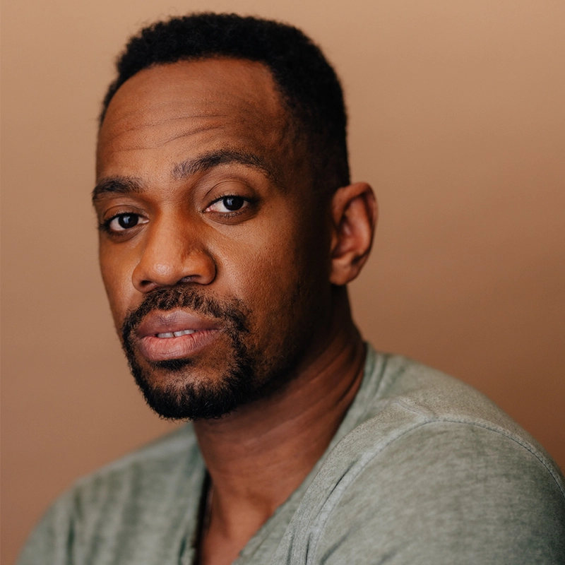 A man with short black hair and a trimmed beard looks into the camera with a neutral expression. He wears a light gray t-shirt. The background is plain and beige, softly lit—photographed in the style of Talia Robinson.