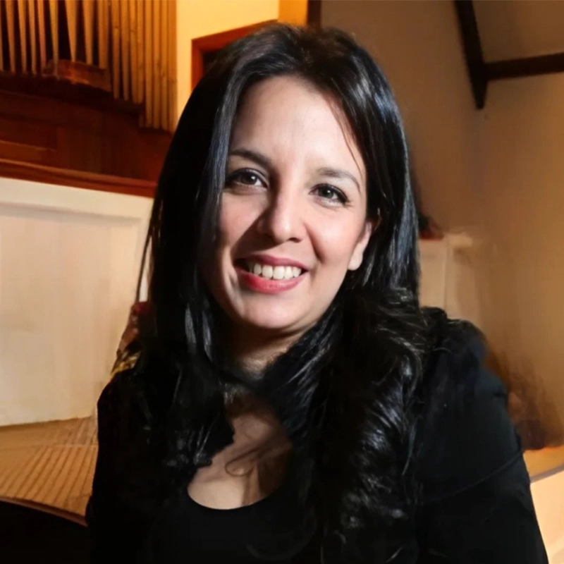A woman with long, dark wavy hair and fair skin smiles at the camera. She is wearing a black top. The warmly lit room with wood paneling and part of a piano or organ creates an inviting backdrop, reminiscent of Khullip Jeung’s musical spaces.