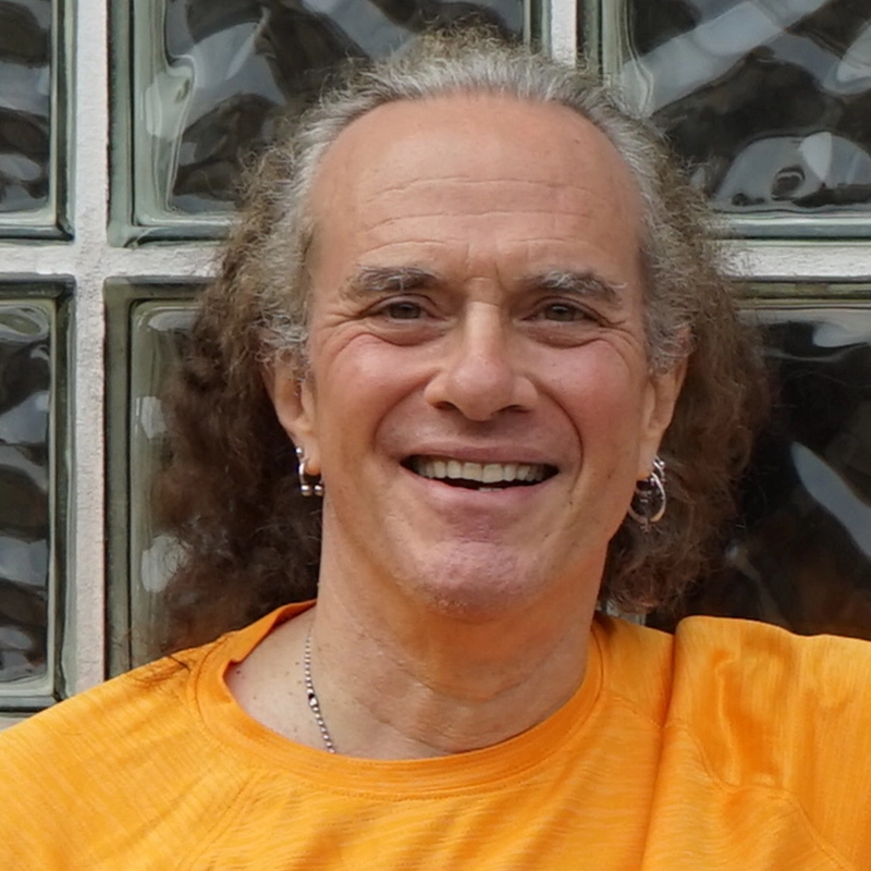 A smiling person with curly long gray hair, wearing hoop earrings and an orange shirt, stands in front of a wall made of square glass blocks. Their expression is friendly and joyful, capturing the warm spirit of Rex Benincasa.