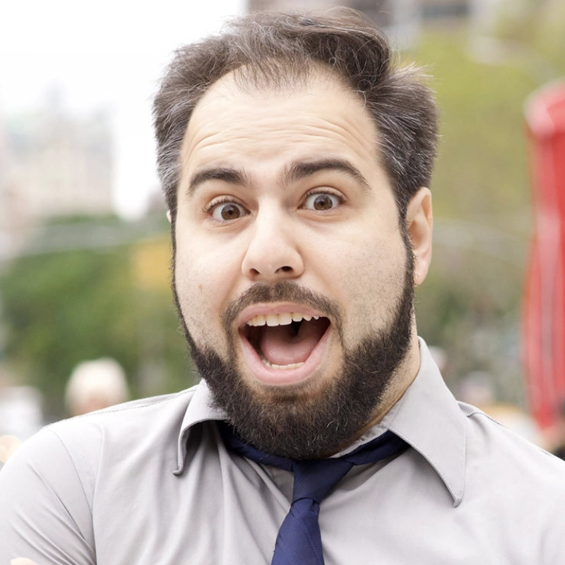 A bearded man with wide eyes and mouth open in surprise or excitement stands outdoors. He wears a light gray shirt and dark blue tie, with his dark hair slightly messy. Blurred greenery and city elements are visible in the background.