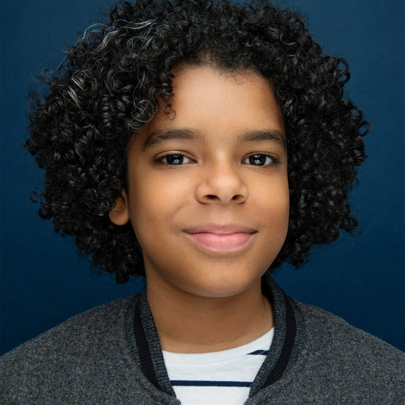 A young boy with medium brown skin and curly black hair smiles gently. He wears a gray jacket with a black collar over a white shirt with blue stripes. The background is solid dark blue. His expression appears calm and friendly.
