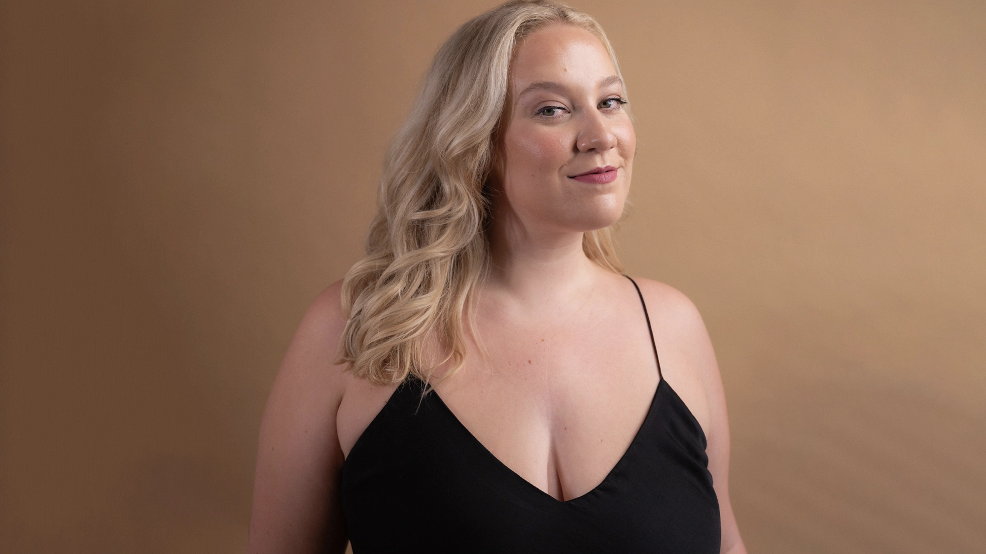 A woman with fair skin and long, wavy blonde hair wears a black sleeveless top with thin straps. She smiles slightly and stands against a warm beige background, channeling the confident spirit of 70s music as she looks at the camera.