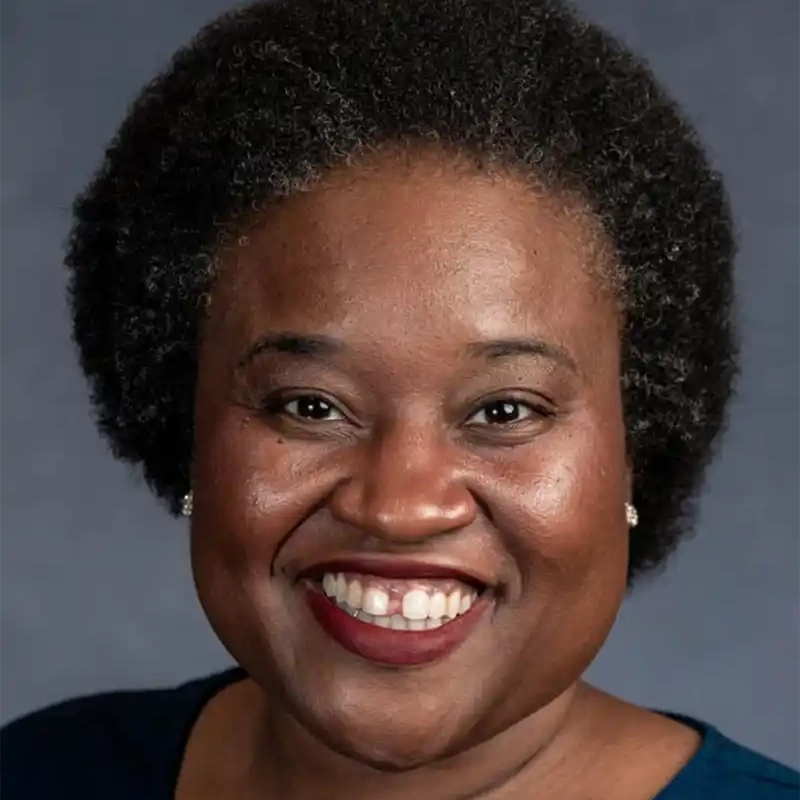 A smiling woman, Shinnerie Jackson, with short, natural curly black hair and medium-dark skin wears pearl stud earrings and a dark blue top. She is facing forward against a plain dark gray background.