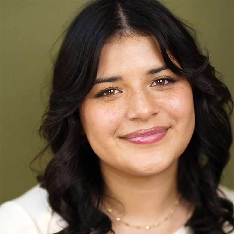 A young woman with medium skin, loose dark wavy hair, and brown eyes smiles gently. She wears a light top and a delicate gold necklace. The background is a plain olive green, creating a warm, inviting atmosphere.